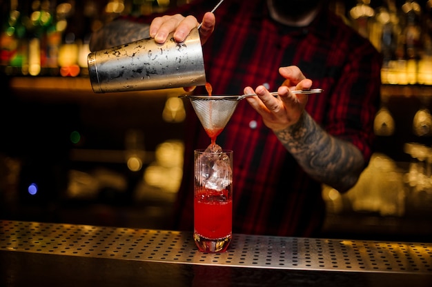 Barman pouring red sweet juicy drink into a cocktail glass using a shaker and strainer