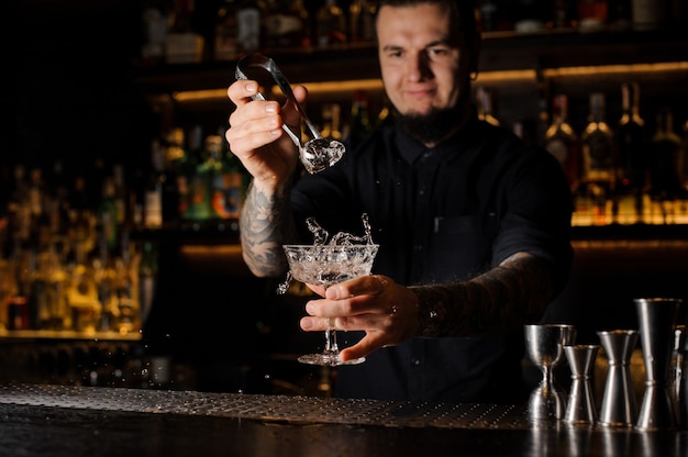 Bartender adding to an alcoholic drink in the glass a big ice cube with tweezers on the bar counter