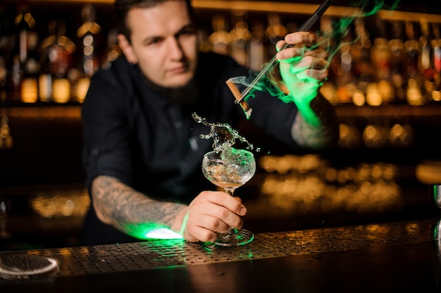 Bartender adding to the cocktail glass with splash with a dried orange aromatic smoked cinnamon with tweezers on the bar counter