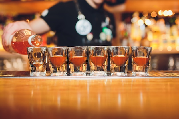 Bartender pouring strong alcoholic drink into small glasses on bar, shots.