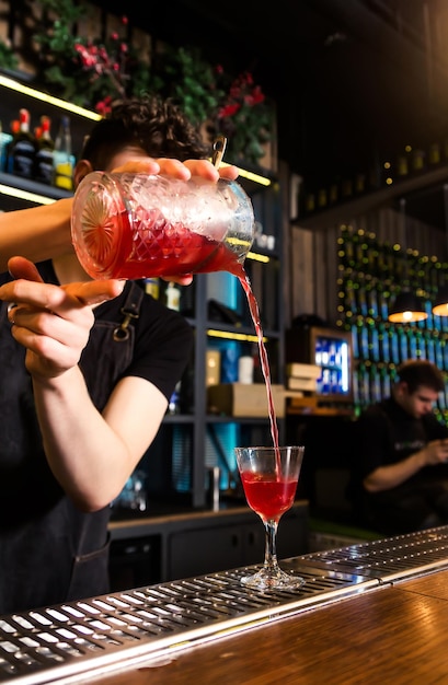 The bartender pours a cocktail from a mixing glass into a cocktail glass