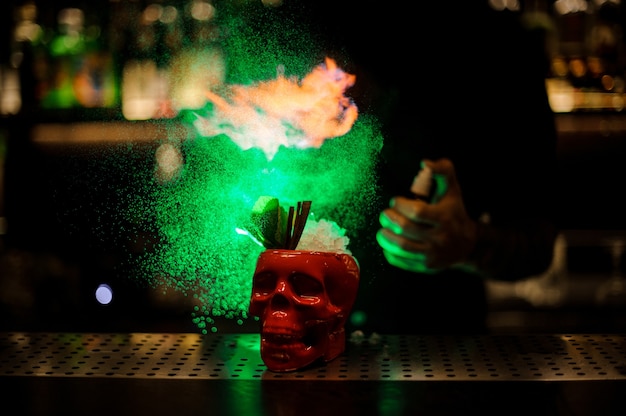 Bartender spraying on the cocktail in the scull red cup from the special vaporizer in the green light on the bar counter.