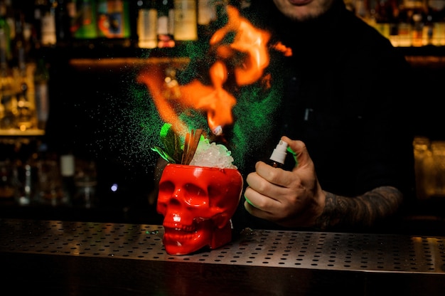 Bartender spraying on the delicious cocktail in the scull cup from the special vaporizer and flame it on the bar counter