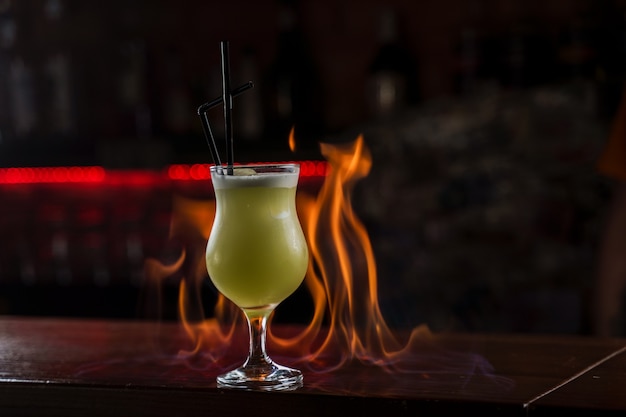 Bartender sprinkles on illuminated glass with bright green cold cocktail on bar counter and makes fire flame over it.