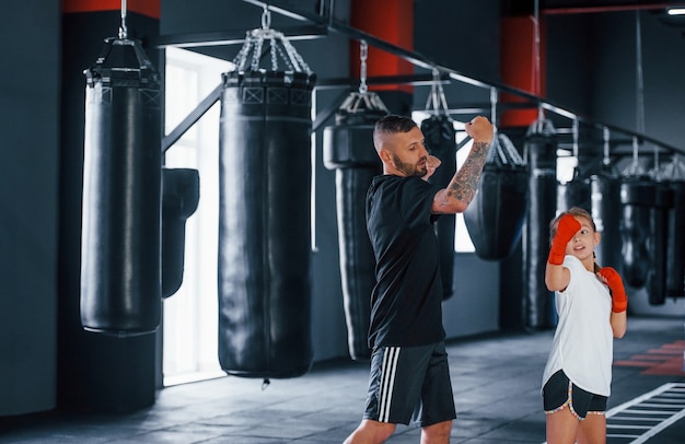 Basic punches. Young tattooed boxing coach teaches cute little girl in the gym.