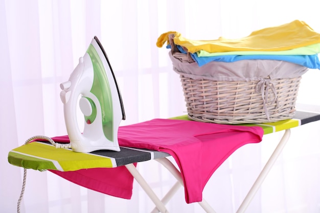 Basket with laundry and ironing board on light home interior background