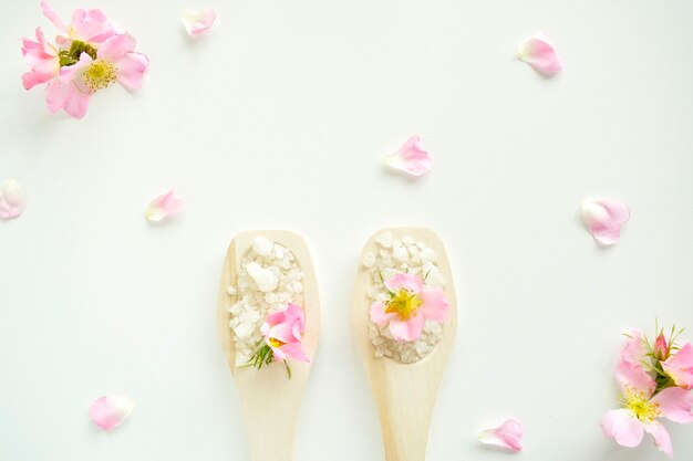 Bath salt in wooden spoons with flowers and rose petals, on a white background. Top view.