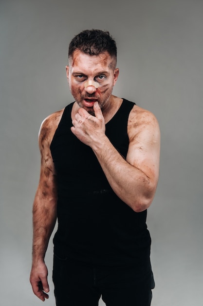 A battered man in a black T shirt stands against a gray wall.