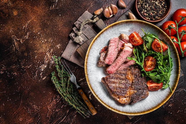 BBQ Grilled rib eye steak, fried rib-eye beef meat on a plate with green salad. Dark background. top view. Copy space.