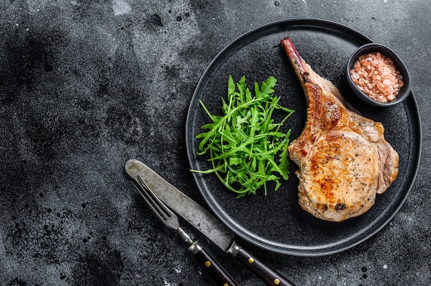 BBQ Grilled tomahawk pork chop meat steak on a marble board. Black background. Top view. Copy space.