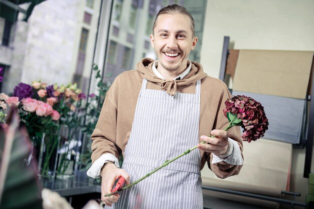 Be happy. Cheerful man keeping smile on his face while holding flower in left hand