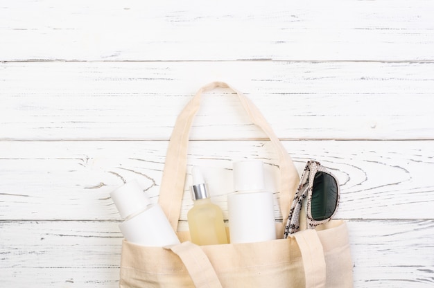 Beach bag with cosmetics, sunscreen, sunglasses on white wooden boards