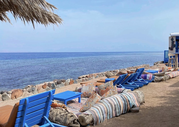 A beach scene with blue chairs and a straw umbrella on the beach.