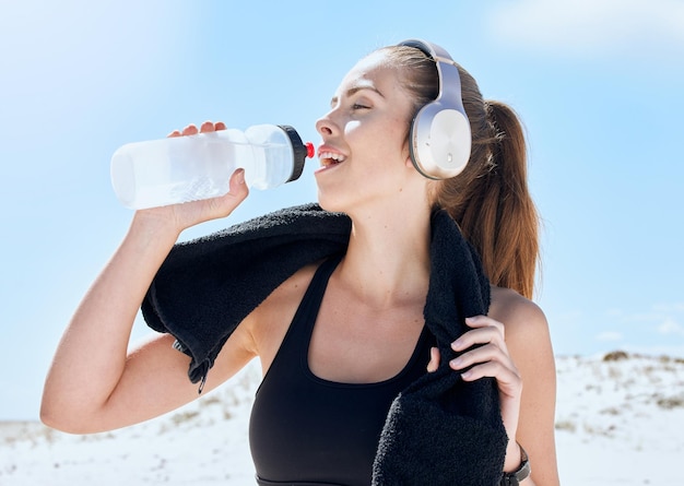 Beach workout and drinking water with headphones music for exercise fitness and training break Wellness relax and sport woman streaming audio and resting body after cardio with water hydration