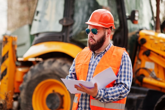 Beard worker man suit construction worker in safety orange helmet, sunglasses against tractor with plan paper at hands.