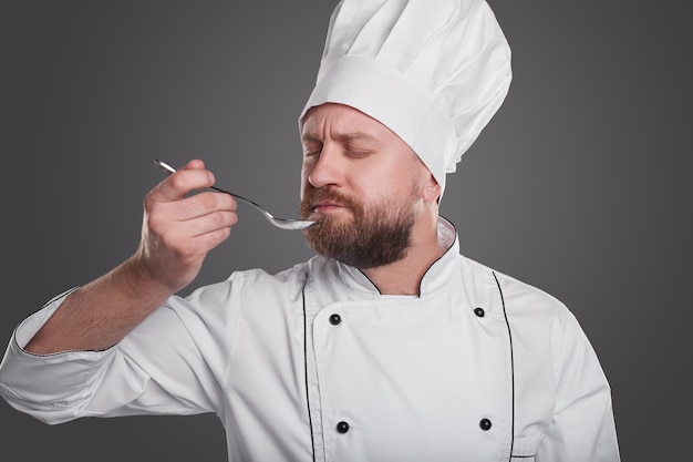 Bearded adult man in chef uniform enjoying smell with closed eyes while tasting food in spoon during work against gray background