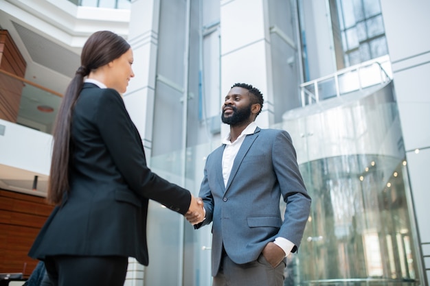 Bearded Afro-American businessman in suit shaking hand of colleague while congratulating her with successful contract