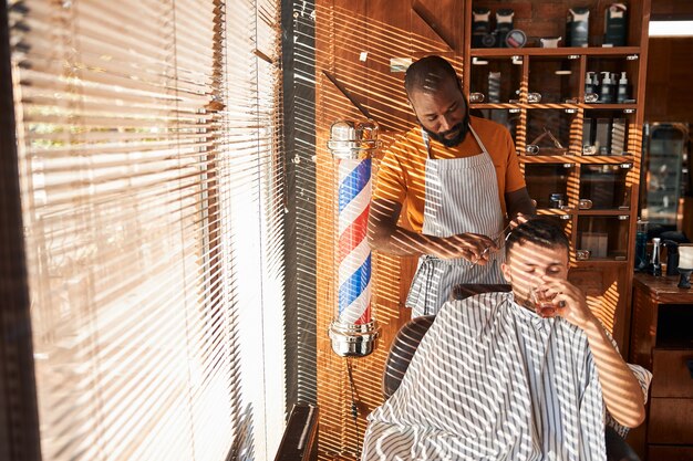 Bearded Afro American male barber cutting client hair with scissors