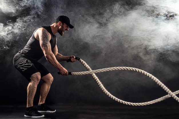 Bearded athletic looking bodybulder work out with battle rope on dark   with smoke. Strength and motivation.