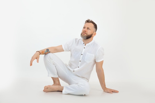 Bearded barefoot thoughtful man with tattoo sitting on the floor in white outfit on white background