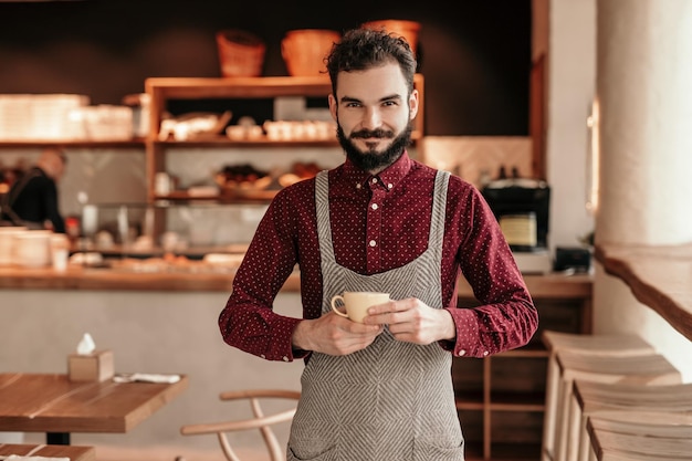 Bearded barista with cup of coffee in cafe