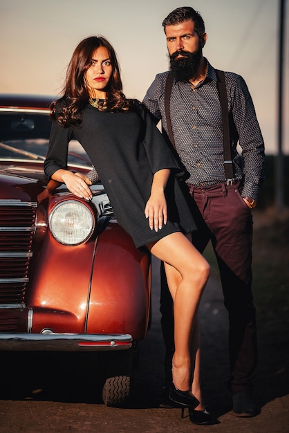 Bearded brunette man in a shirt and trousers with suspenders hugs a slender young girl with long hair in a black short dress near a brown retro car on a dark field background