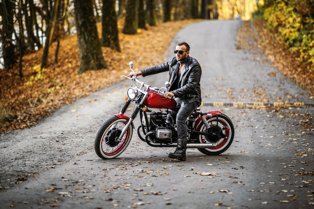 Bearded brutal man in sunglasses and leather jacket sitting on a motorcycle on the road in the forest