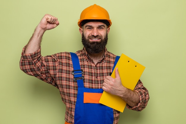 Bearded builder man in construction uniform and safety helmet holding clipboard looking happy and excited raising fist