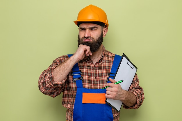 Bearded builder man in construction uniform and safety helmet holding clipboard with pen looking aside puzzled standing over green background