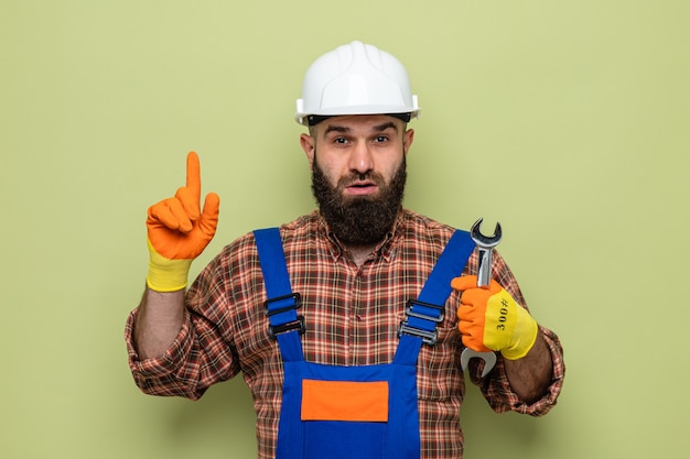 Bearded builder man in construction uniform and safety helmet wearing rubber gloves holding wrench looking surprised pointing with index finger to the side