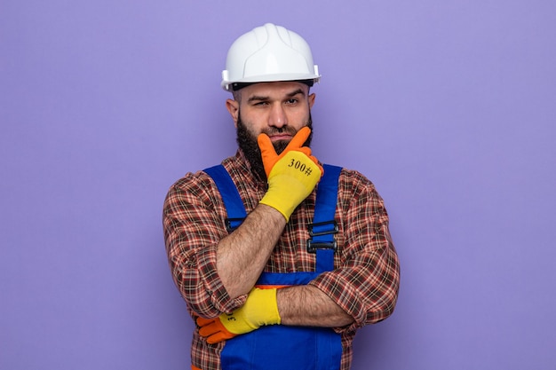 Bearded builder man in construction uniform and safety helmet wearing rubber gloves looking with hand on his chin thinking