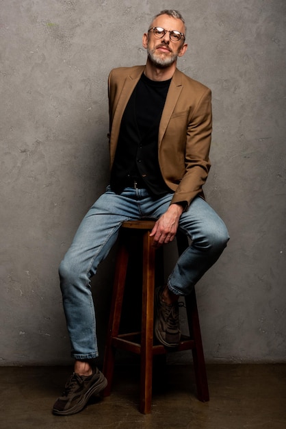 Bearded businessman in glasses sitting on stool and looking at camera on grey