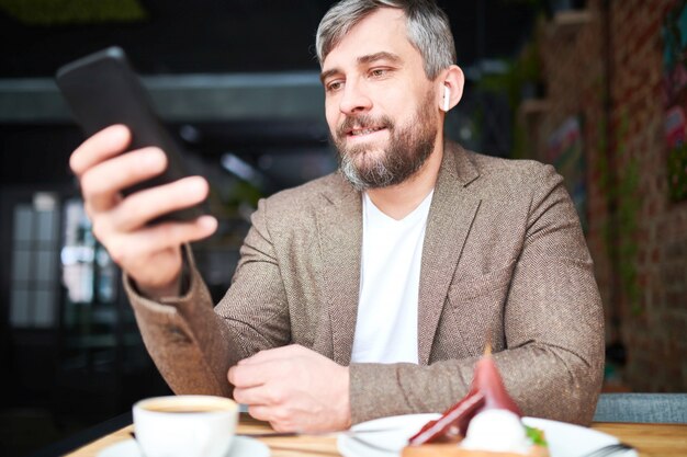 Bearded businessman in smart casual sitting by table in cafe and messaging in his smartphone after work