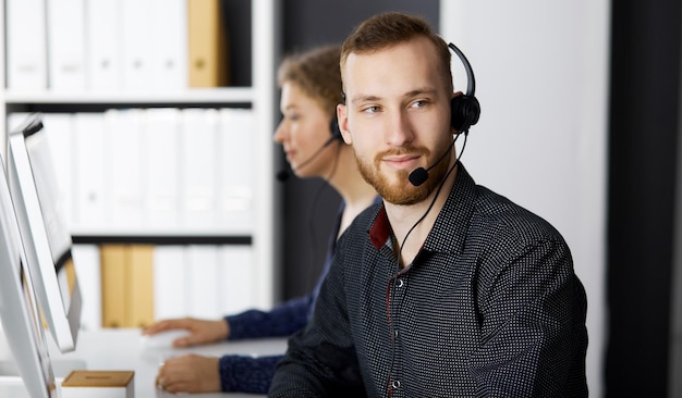 Bearded businessman talking by headset near his female colleague while sitting in modern office. Diverse people group in call center. Telemarketing and customer service