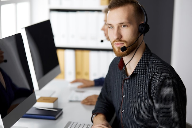 Bearded businessman talking by headset near his female colleague while sitting in modern office. Diverse people group in call center. Telemarketing and customer service.