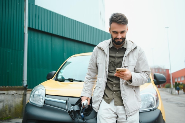 Bearded caucasian man standing near an electric car that is charging and making time adjustments on a smartphone