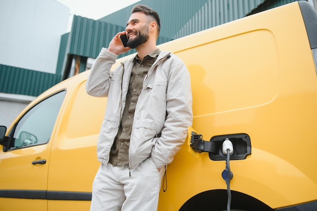 Bearded caucasian man standing near an electric car that is charging and making time adjustments on a smartphone