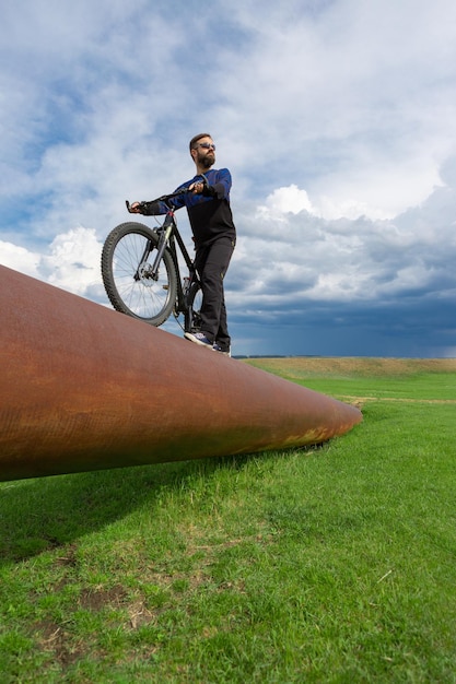 Bearded cyclist on a mountain bike on a rusty pipe green grass blue sky