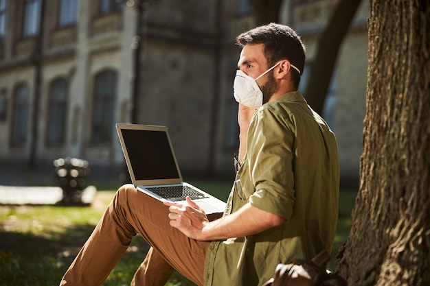 Bearded dark-haired young male in a protective mask and casual clothes sitting under a tree