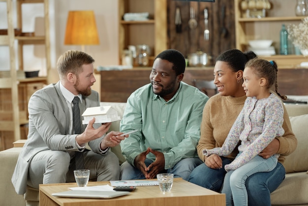 Bearded elegant adviser with mockup house consulting adult ethnic couple with kid on new real estate