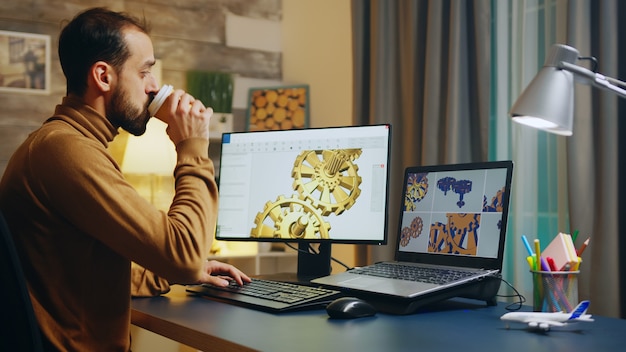 Bearded engineer taking a sip of coffee while working on computer. Gears production.