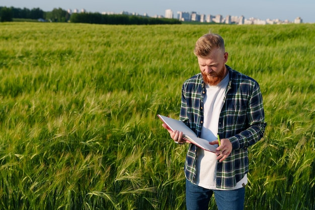 A bearded farmer with a notebook stands in a wheat field against the backdrop of a megalopolis Preparing the field for a good ecological harvest