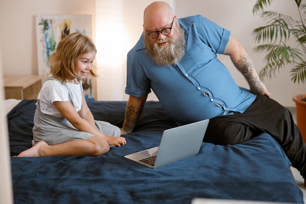 Bearded father and little girl watch moovie lesson via modern laptop sitting on bed