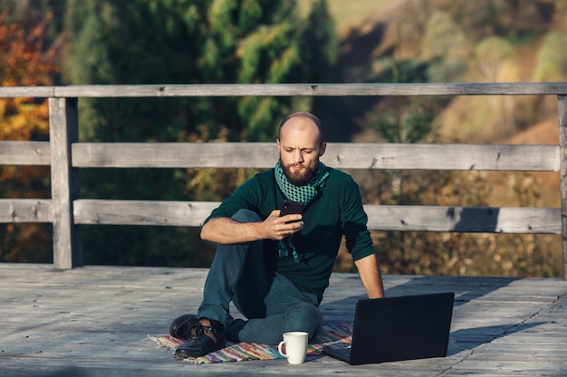 Bearded freelancer man sitting on terrace working on laptop and talking on phone