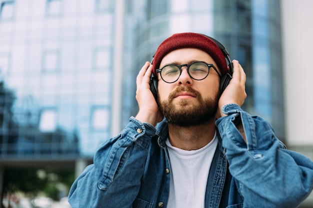 Bearded hipster male enjoys music with closed eyes in headphones outdoors in the city. Room for text.