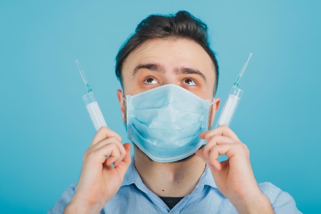 bearded male doctor in mask holding two syringes in his hands on blue background