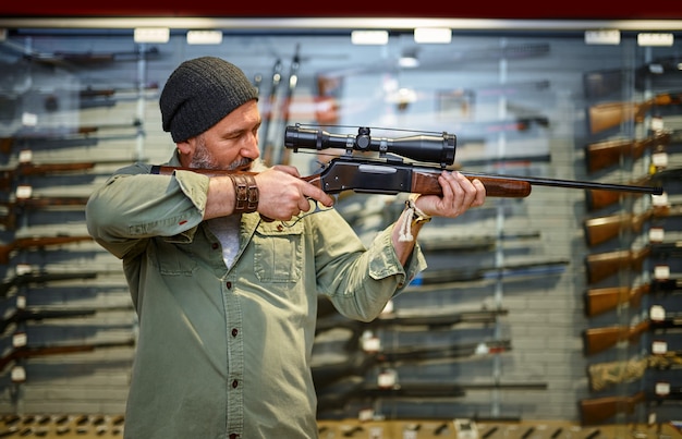 Bearded male hunter buying hunting rifle with optical sight in gun store. Weapon shop interior, ammo and ammunition assortment, firearms choice, shooting hobby