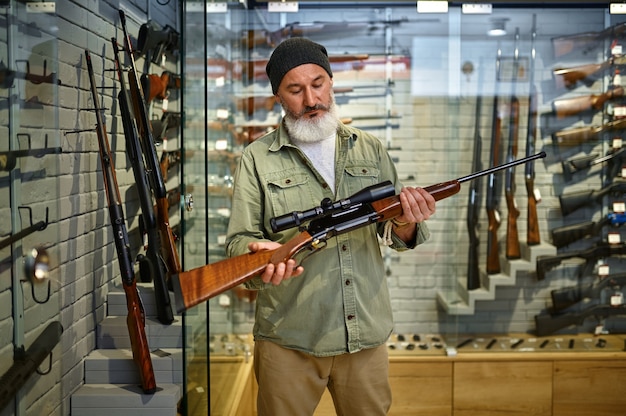 Bearded male hunter holds hunting rifle with optical sight in gun store. Weapon shop interior, ammo and ammunition assortment, firearms choice, shooting hobby and lifestyle