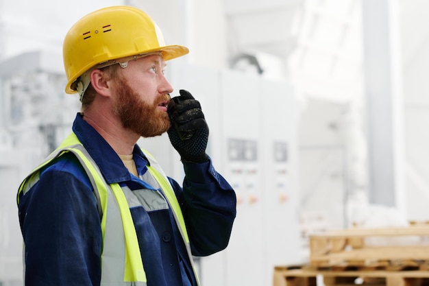 Bearded male worker in uniform hardhat and black gloves using walkietalkie