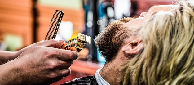 Bearded man in barbershop Man visiting hairstylist in barbershop Barber works with a beard clipper Hipster client getting haircut Hands of a hairdresser with a beard clipper closeup